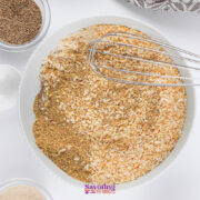 A mixing bowl filled with dried minced onions, spices, and Onion Soup Mix sits beside a whisk, two measuring spoons, and small bowls of seasonings on a white surface.