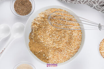 A mixing bowl filled with dried minced onions, spices, and Onion Soup Mix sits beside a whisk, two measuring spoons, and small bowls of seasonings on a white surface.