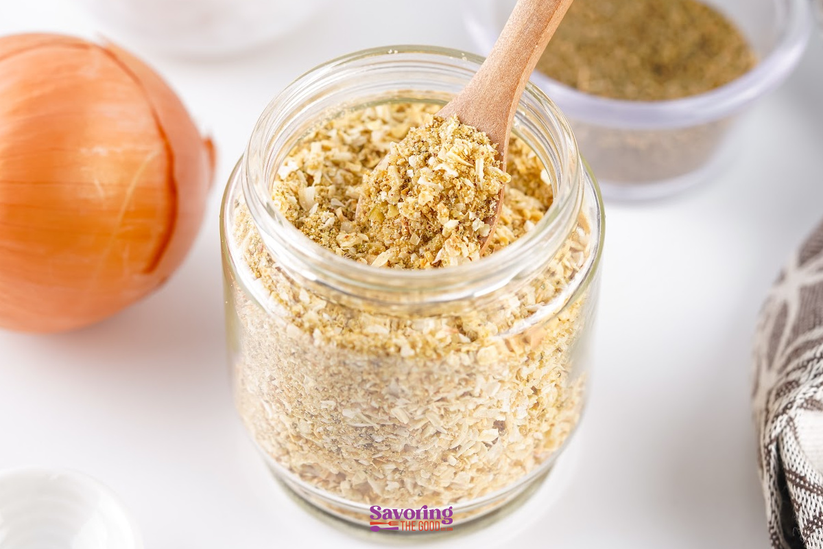 A glass jar filled with Onion Soup Mix sits on a white surface, with a wooden spoon inside the jar and an unpeeled onion nearby.