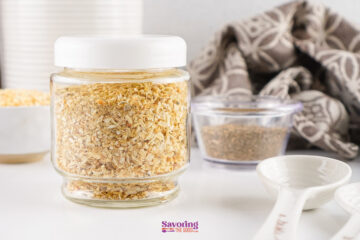 A glass jar filled with dried minced onions, perfect for making onion soup mix, sits on a white surface next to measuring spoons, a small bowl of spices, and a patterned cloth.