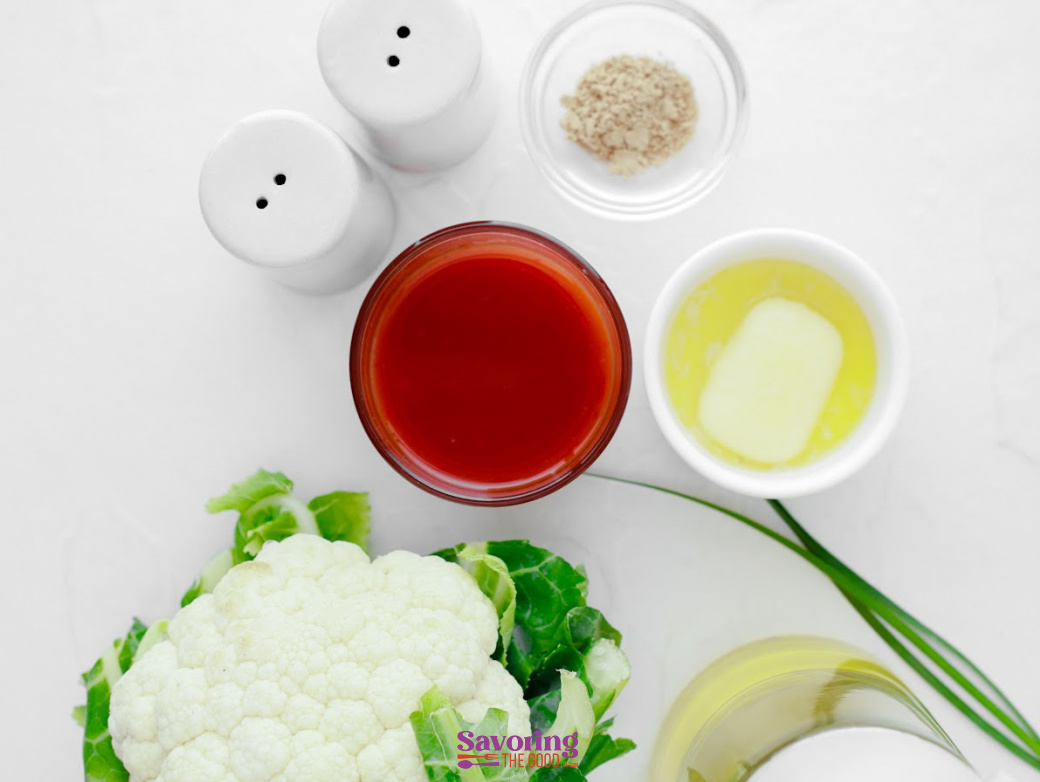 Overhead view of ingredients for Buffalo Cauliflower Bites: cauliflower, a bowl of hot sauce, melted butter, salt, pepper, garlic powder, and chives arranged on a white surface.