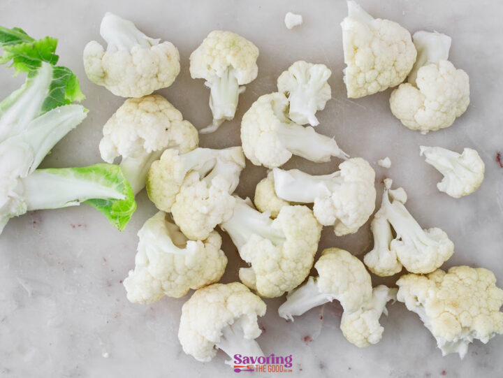 Raw cauliflower florets and a few leaves are scattered on a light-colored surface.