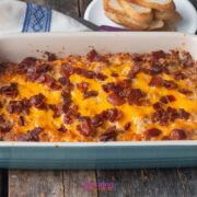 A baked casserole topped with melted cheddar cheese and pieces of bacon is shown in a rectangular dish on a wooden table. Toasted bread slices and a bowl of caramelized onion dip are in the background.