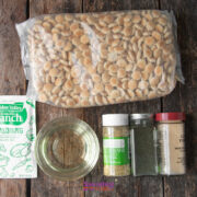 A bag of oyster crackers, a packet of ranch seasoning, a bowl of oil, and three spice jars are arranged on a wooden surface.