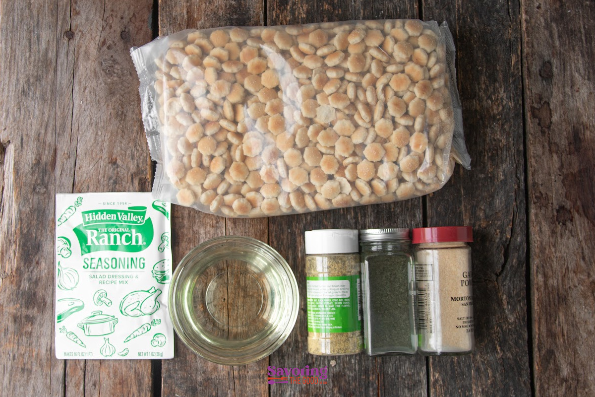 A bag of oyster crackers, a packet of ranch seasoning, a bowl of oil, and three spice jars are arranged on a wooden surface.
