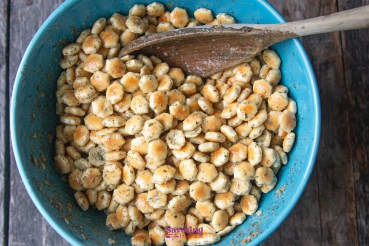 A blue bowl filled with seasoned oyster crackers being mixed with a wooden spoon on a wooden surface.