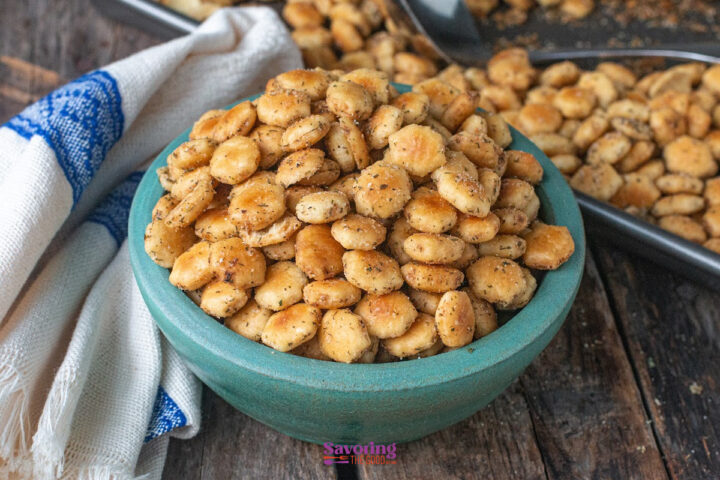 A teal bowl filled with seasoned oyster crackers sits on a wooden table, with more crackers scattered on a baking tray in the background.