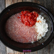 A slow cooker filled with ground meat, diced tomatoes, and chopped onions sits on a wooden surface, ingredients uncooked and unmixed.