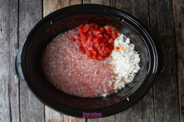 A slow cooker filled with ground meat, diced tomatoes, and chopped onions sits on a wooden surface, ingredients uncooked and unmixed.