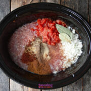 A slow cooker filled with ground meat, diced onions, canned tomatoes, bay leaves, and various spices on a wooden surface.