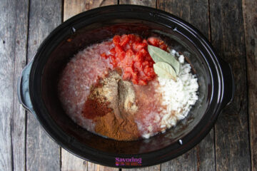 A slow cooker filled with ground meat, diced onions, canned tomatoes, bay leaves, and various spices on a wooden surface.