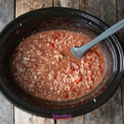 A slow cooker filled with a mixture of ground meat, chopped onions, tomatoes, and sauce, with a gray spoon resting inside.