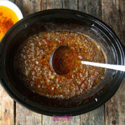 A slow cooker filled with brown, chunky broth sits on a wooden surface, with a black ladle resting inside and a bowl of orange liquid nearby.