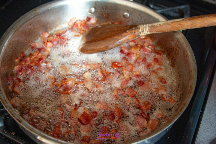 Chopped bacon pieces frying in a stainless steel skillet with bubbling grease, stirred by a wooden spoon on a stovetop.