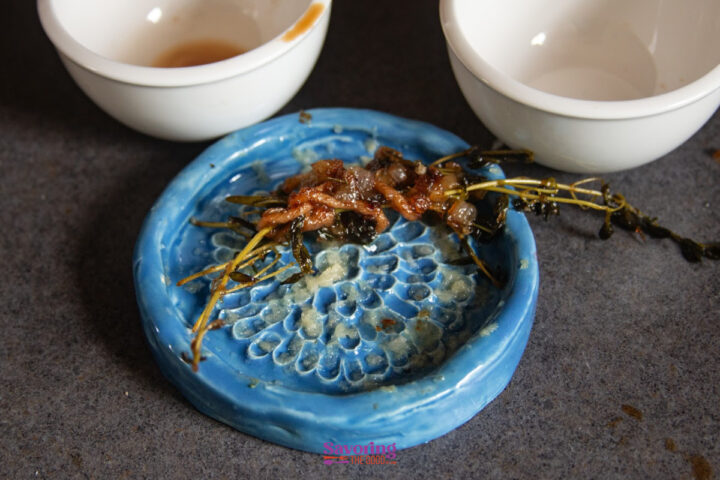 A small portion of cooked greens and possibly meat sits on a blue ceramic dish, with two white bowls in the background on a dark surface.