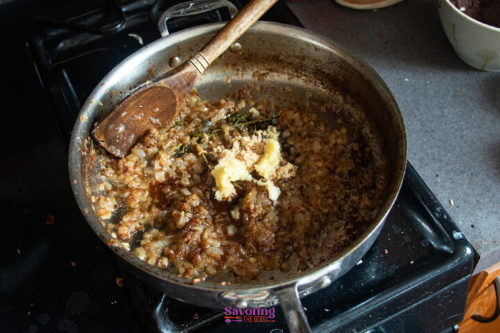 Chopped onions sautéing in a pan with brown sugar, butter, and herbs, stirred with a wooden spoon on a stovetop.