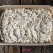 A beige rectangular baking dish filled with an unbaked, creamy caramelized onion dip mixture-speckled with visible bits of vegetables and possibly bacon-rests on a wooden surface.