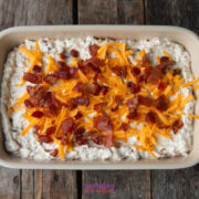 A rectangular baking dish filled with a creamy caramelized onion dip mixture, topped with shredded cheddar cheese and chopped cooked bacon, sits on a wooden surface.