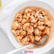 A white bowl filled with cauliflower florets coated in a reddish-brown sauce, with a spoon resting in the bowl, on a white surface with a white napkin.