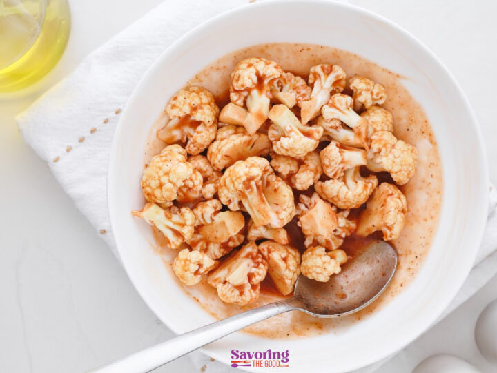 A white bowl filled with cauliflower florets coated in a reddish-brown sauce, with a spoon resting in the bowl, on a white surface with a white napkin.