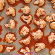 Buffalo Cauliflower Bites featuring raw cauliflower florets coated in a spicy red sauce are evenly spread on a parchment-lined baking sheet, ready to bake.