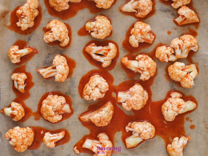 Buffalo Cauliflower Bites featuring raw cauliflower florets coated in a spicy red sauce are evenly spread on a parchment-lined baking sheet, ready to bake.
