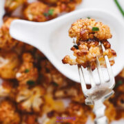 A fork holds a roasted Buffalo Cauliflower Bite above a plate with more pieces and a white condiment spoon.