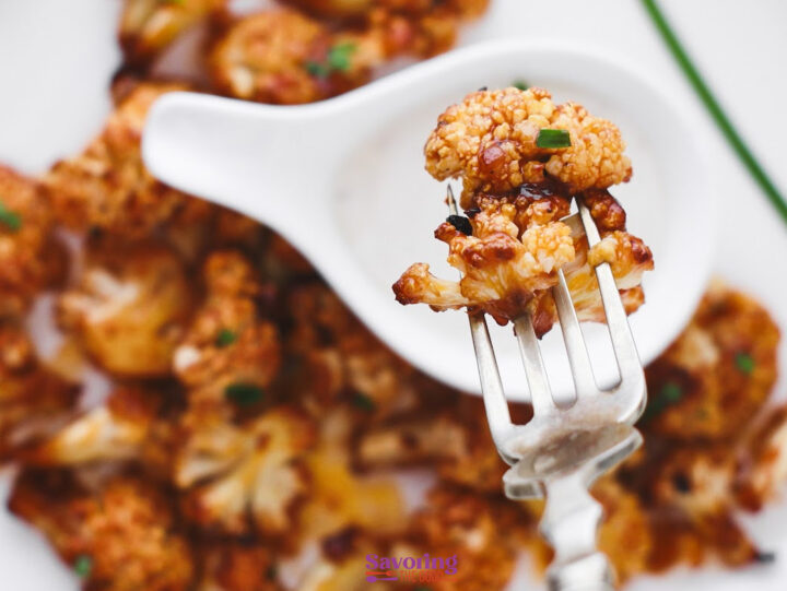 A fork holds a roasted Buffalo Cauliflower Bite above a plate with more pieces and a white condiment spoon.