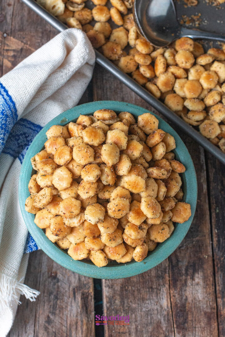 A bowl of seasoned oyster crackers sits on a wooden table next to a tray of more crackers and a white towel with a blue stripe.