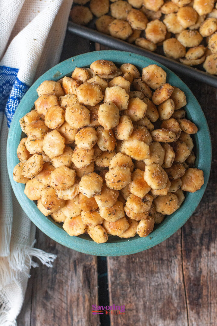 A bowl filled with seasoned oyster crackers sits on a wooden surface next to a tray of more crackers and a folded cloth.