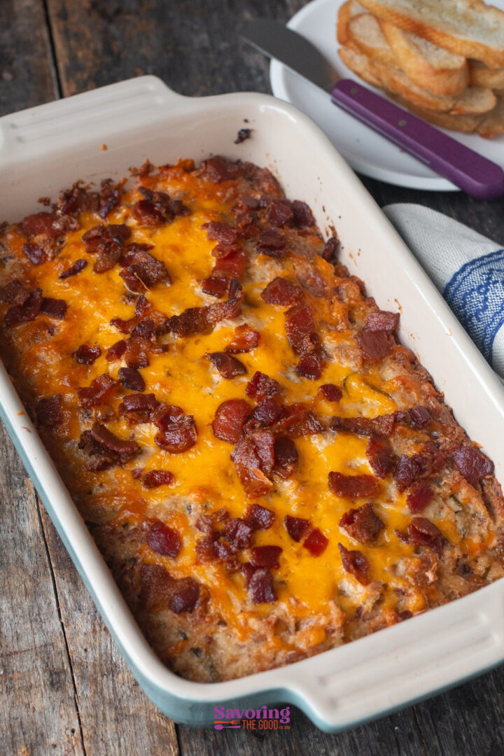 A baked casserole topped with melted cheddar cheese and crumbled bacon sits in a rectangular dish on a wooden table, reminiscent of caramelized onion dip, with a plate of toast and a knife in the background.