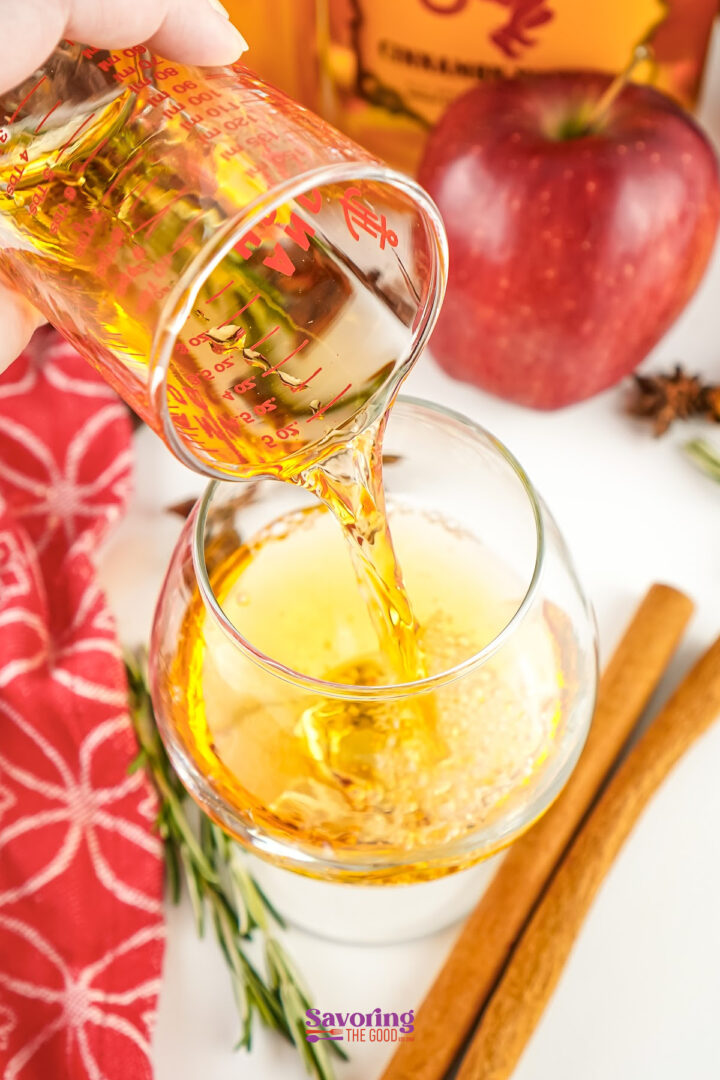 A hand pours a golden liquid from a measuring cup into a glass. Nearby, an apple, cinnamon sticks, and a red patterned cloth hint at cozy apple cider with Fireball.