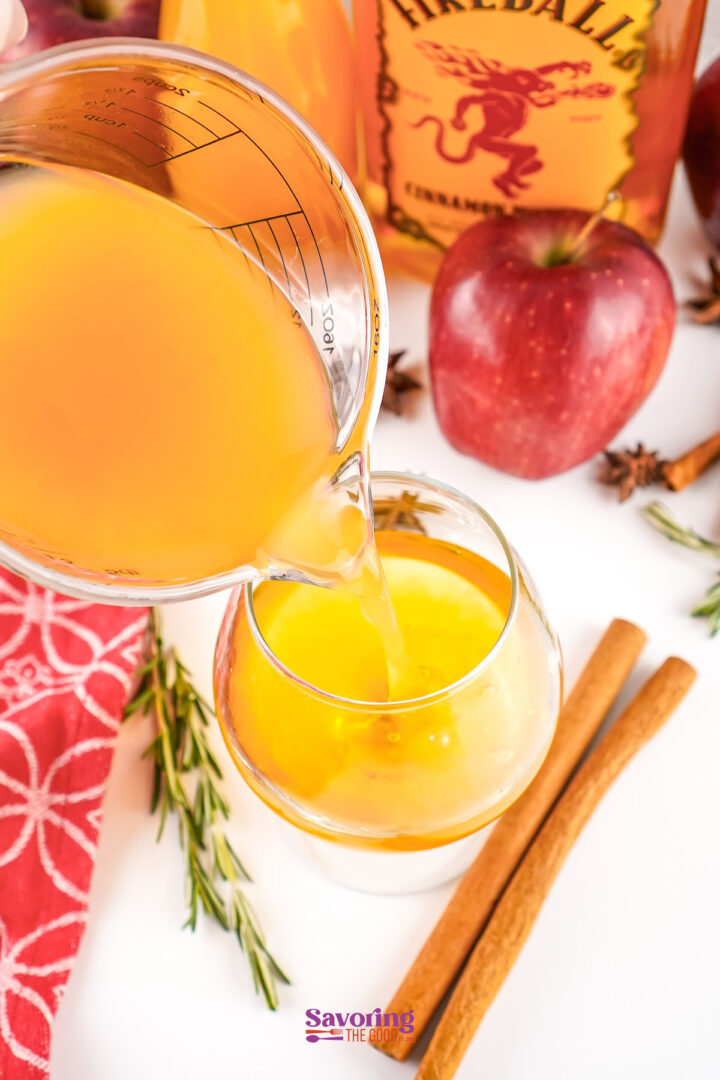 A measuring cup pours a yellow-orange liquid into a glass, surrounded by apples, cinnamon sticks, and a bottle of Fireball whiskey-perfect for making apple cider with Fireball.