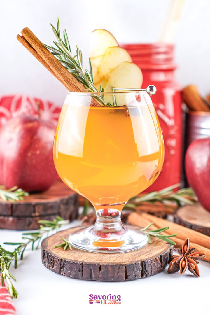 A glass of apple cider with Fireball garnished with apple slices, a cinnamon stick, and rosemary sits on a wooden coaster, surrounded by apples and spices in the background.