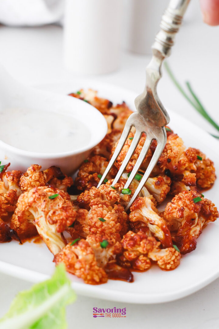 A fork holds a piece of Buffalo Cauliflower Bites on a plate with ranch dipping sauce and chopped chives sprinkled on top.