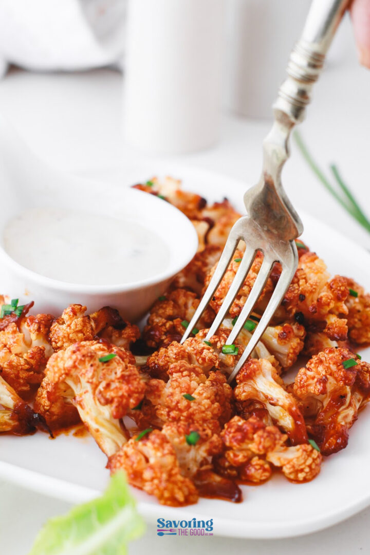 A fork picks up a piece of buffalo cauliflower from a plate next to a small bowl of white dipping sauce.