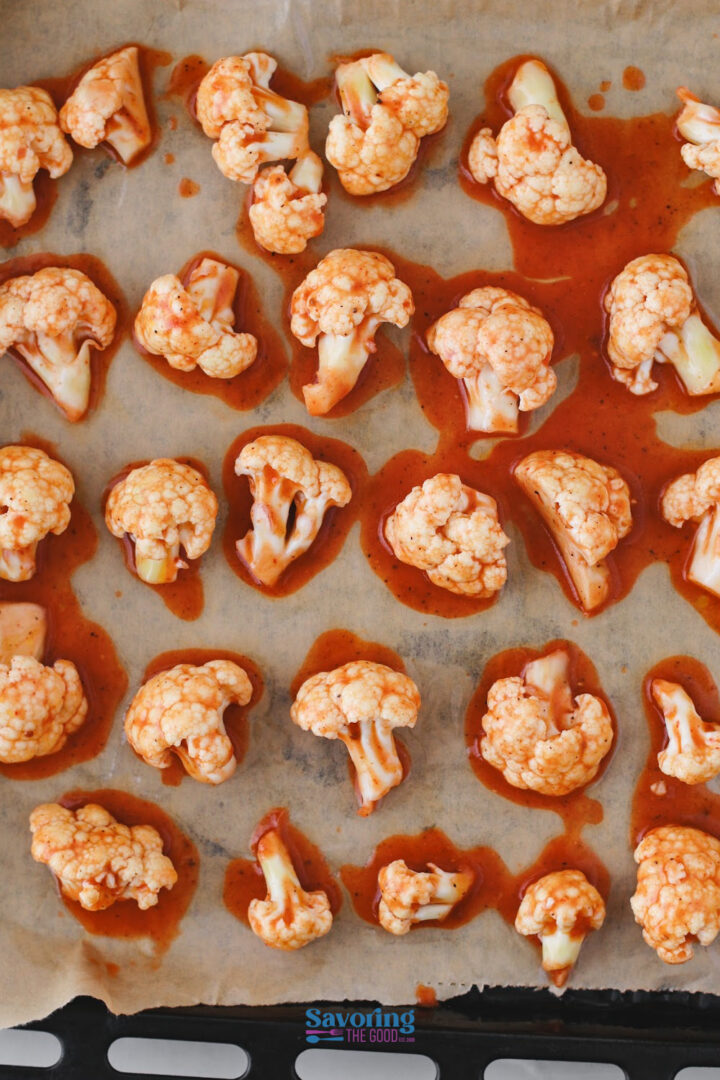 Raw cauliflower florets coated in a red sauce arranged on parchment paper on a baking tray.