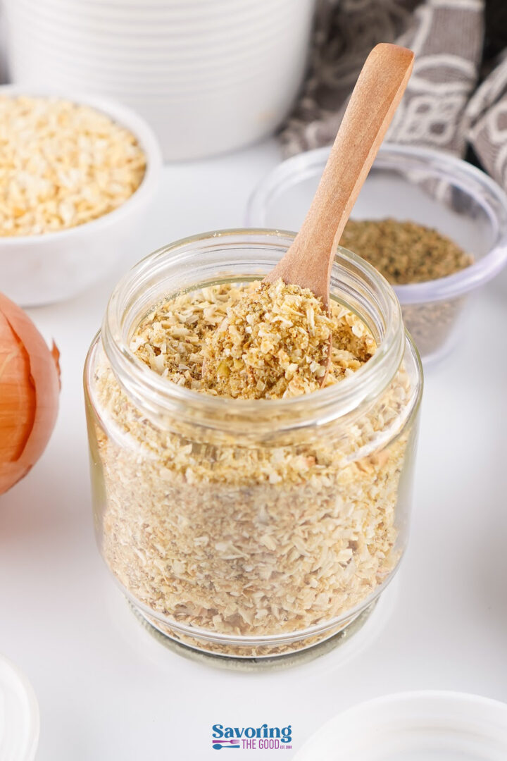 A glass jar filled with dried minced onions, with a wooden spoon inside. Other spices and an onion are visible in the background.