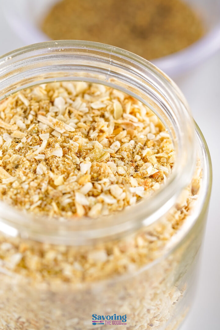 A close-up of a glass jar filled with a dry seasoning blend containing dried onion flakes and various spices.