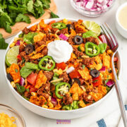 A bowl of taco salad with ground beef, lettuce, tomatoes, black olives, jalapeños, corn, shredded cheese, crushed tortilla chips, red onions, lime wedges, and a dollop of sour cream.