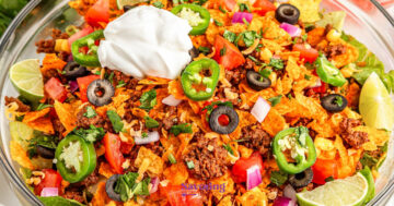 A bowl of taco salad topped with ground beef, tomato, jalapeños, black olives, red onion, shredded cheese, cilantro, lime wedges, and a dollop of sour cream.