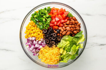 A glass bowl filled with separate sections of lettuce, shredded cheese, black olives, red onion, corn, green bell pepper, diced tomato, and pinto beans on a white surface.