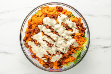 A glass bowl filled with lettuce, orange tortilla chips, seasoned ground beef, and creamy dressing arranged in layers.