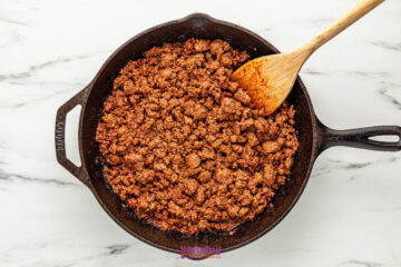 Cooked ground beef with seasoning in a cast iron skillet, stirred with a wooden spoon on a white marble surface.