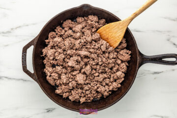 Cooked ground beef in a cast iron skillet with a wooden spatula resting on the edge, on a white marble surface.