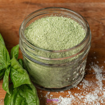 A small glass jar filled with green basil salt sits on a wooden surface next to fresh basil leaves and scattered salt.