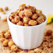 A white cup filled with roasted chickpeas coated in seasoning and grated cheese, with some chickpeas scattered around on a brown surface.
