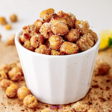 A white cup filled with roasted chickpeas coated in seasoning and grated cheese, with some chickpeas scattered around on a brown surface.