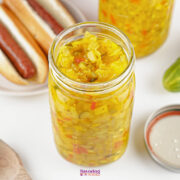 A jar of homemade sweet pickle relish sits on a white surface next to hot dogs, an open jar lid, and a wooden spoon.