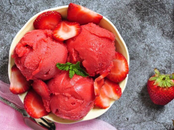 A bowl filled with three scoops of strawberry sorbet, garnished with fresh strawberry slices and a sprig of mint, set on a gray surface.